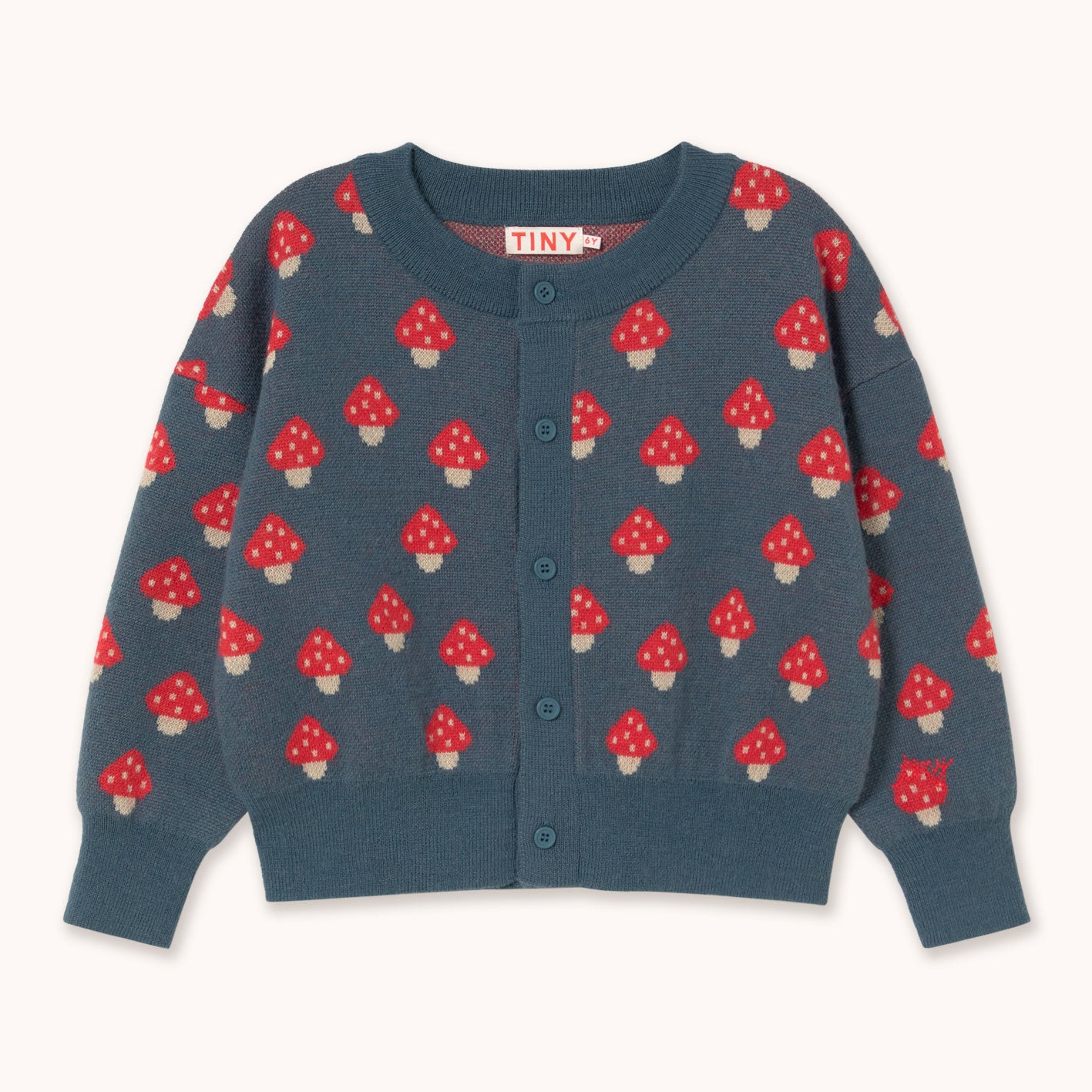 A children's long-sleeve cardigan sweater by designer Tiny Cottons in a slate blue-gray color, featuring an all-over pattern of red and white mushrooms. The cardigan has a button-up front and ribbed collar, cuffs, and hem.