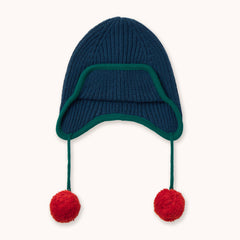 A navy blue ribbed knit winter hat by designer Tiny Cottons with ear flaps, a dark green trim, and two red pom-poms hanging from the sides, shown against a plain off-white background.