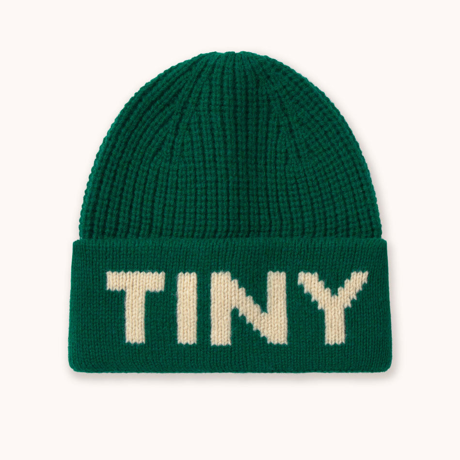 A dark green ribbed knit beanie hat by designer Tiny Cottons with the word 'TINY' knitted in cream-colored letters across the folded cuff, shown against a plain off-white background.