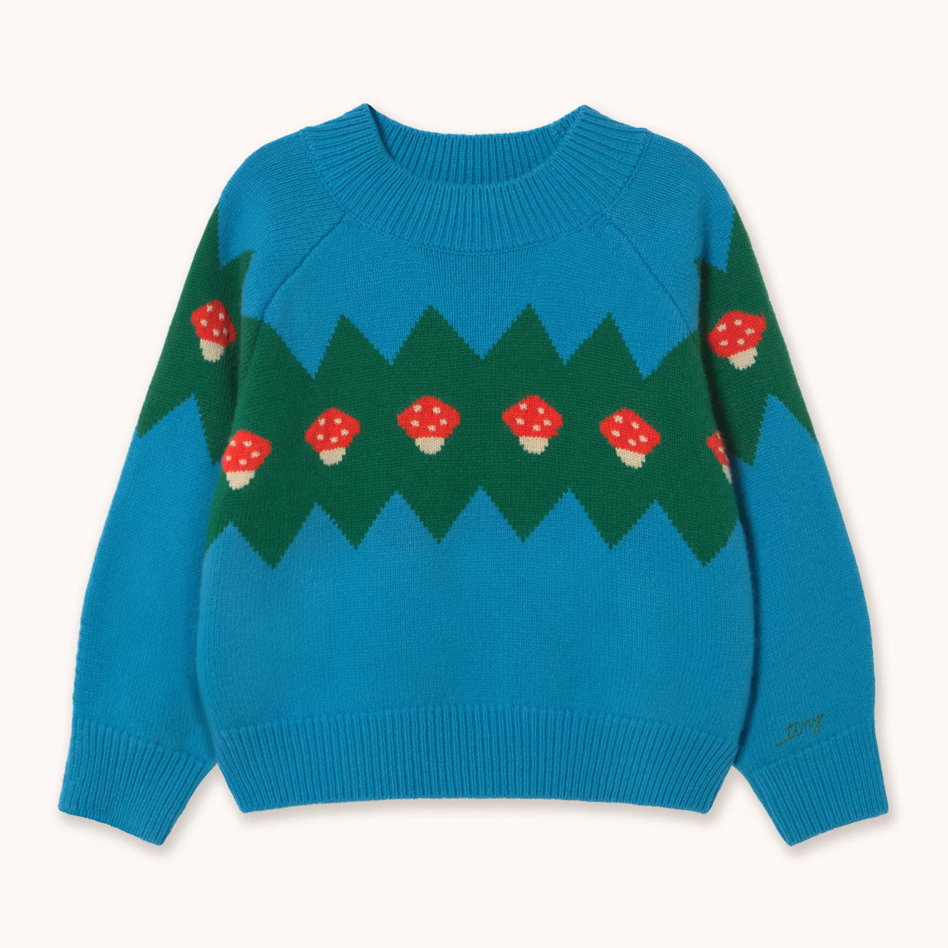 A bright blue and green knit sweater by designer Tiny Cottons with red and white mushroom motifs, laid flat against a light cream background.