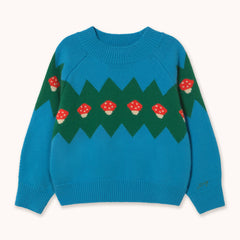 A bright blue and green knit sweater by designer Tiny Cottons with red and white mushroom motifs, laid flat against a light cream background.