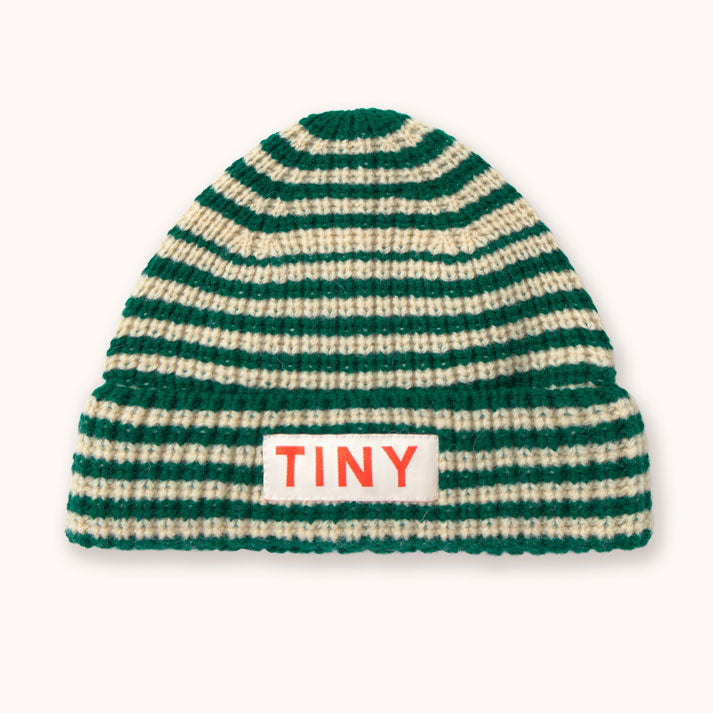 A green and cream striped knit beanie hat by designer Tiny Cottons with a white rectangular patch on the front cuff featuring the word 'TINY' in orange-red letters, shown against a plain off-white background. 