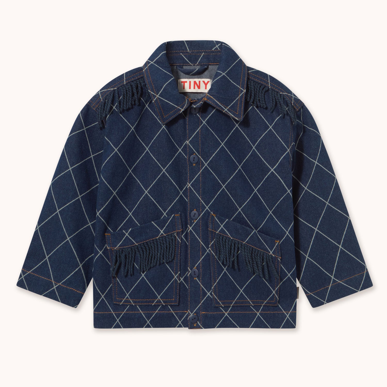 A navy blue denim jacket with a white argyle pattern and fringe details on the pockets and shoulders by designer Tiny Cottons shown flat against a light cream background. The brand label inside the collar reads 'TINY'.
