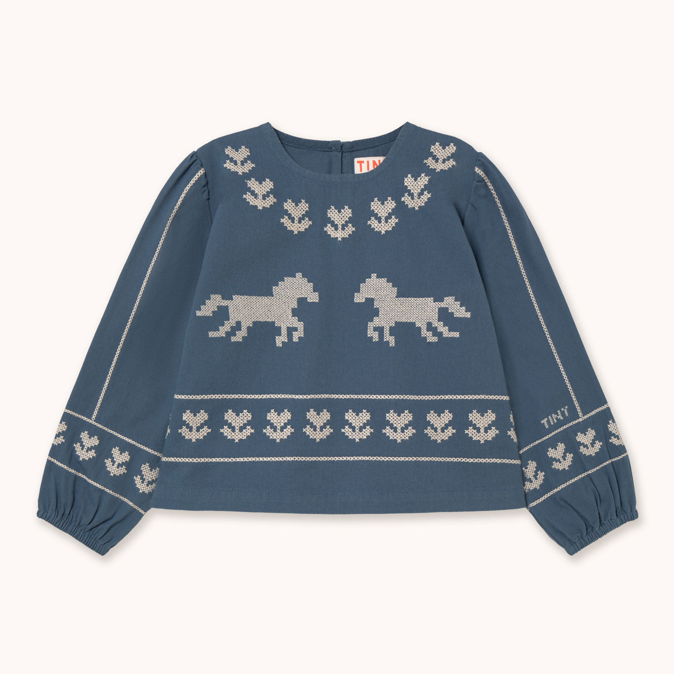 A blue knit sweater with cream-colored pixelated horse motifs and decorative borders by designer Tiny Cottons, laid flat on a light cream background. The brand name 'THAY' is visible on the sleeve and inner neck label.