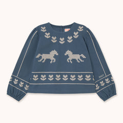 A blue knit sweater with cream-colored pixelated horse motifs and decorative borders by designer Tiny Cottons, laid flat on a light cream background. The brand name 'THAY' is visible on the sleeve and inner neck label.