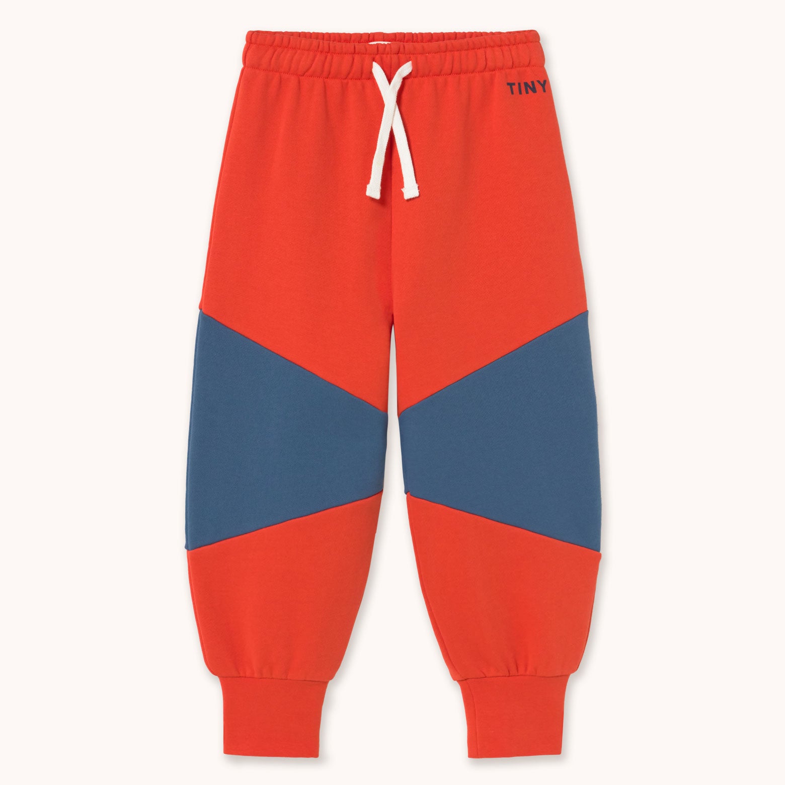 A pair of children's jogger sweatpants in a vibrant red-orange color by designer Tiny Cottons, featuring a geometric color-blocked design with navy blue panels on the thighs and lower legs, a white drawstring, and ribbed cuffs. The brand name 'TINY' is printed in navy on the upper right hip.