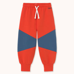 A pair of children's jogger sweatpants in a vibrant red-orange color by designer Tiny Cottons, featuring a geometric color-blocked design with navy blue panels on the thighs and lower legs, a white drawstring, and ribbed cuffs. The brand name 'TINY' is printed in navy on the upper right hip.