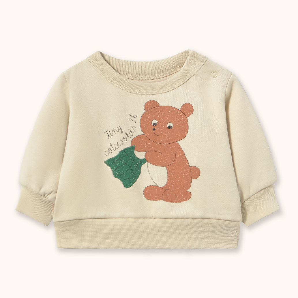 A cream-colored long-sleeve baby sweatshirt by designer Tiny Cottons featuring a brown teddy bear graphic holding a green blanket, with the text 'tiny cotswolds 26' printed on the front.