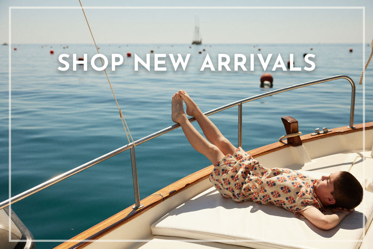 Boy wearing patterned shorts and top by designer Bebe Organic lying on a boat with 'Shop New Arrivals' text overlay